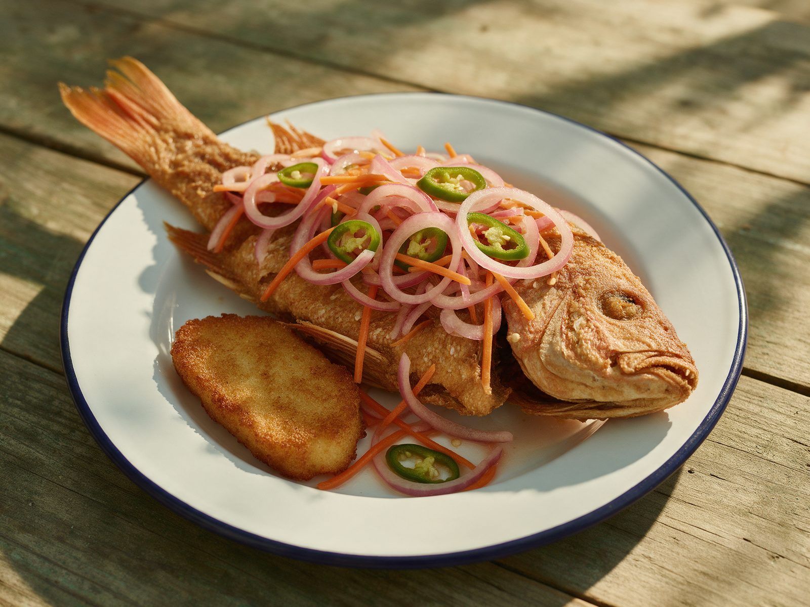 A whole fried snapper topped with pickled onion, carrot and pepper, with a piece of fried bammy.